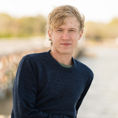 Young adult man standing outdoors near water, looking at the camera.