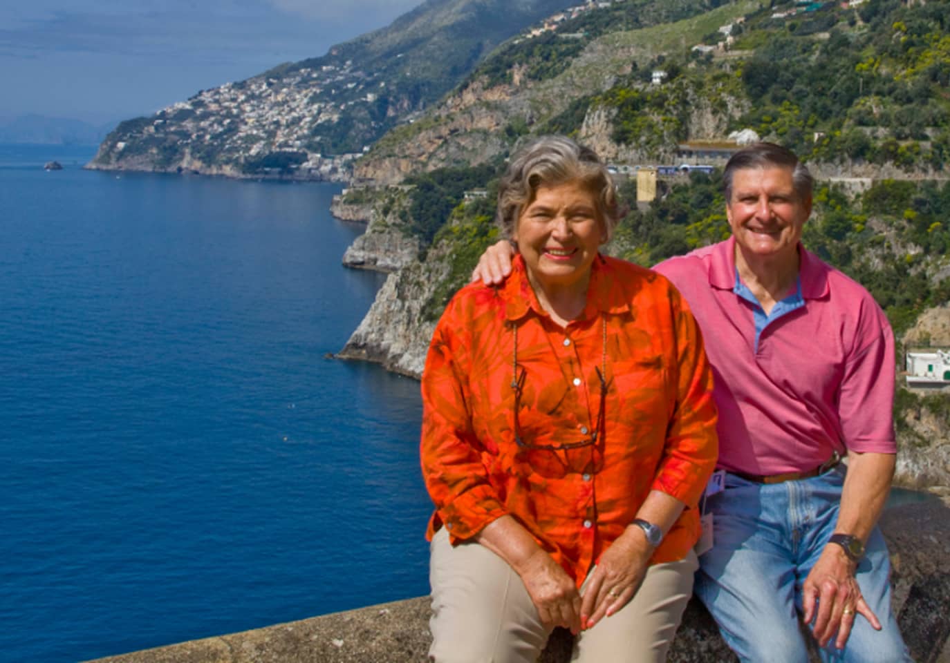 Elderly couple smiling, sitting on a ledge with a scenic coastal backdrop.