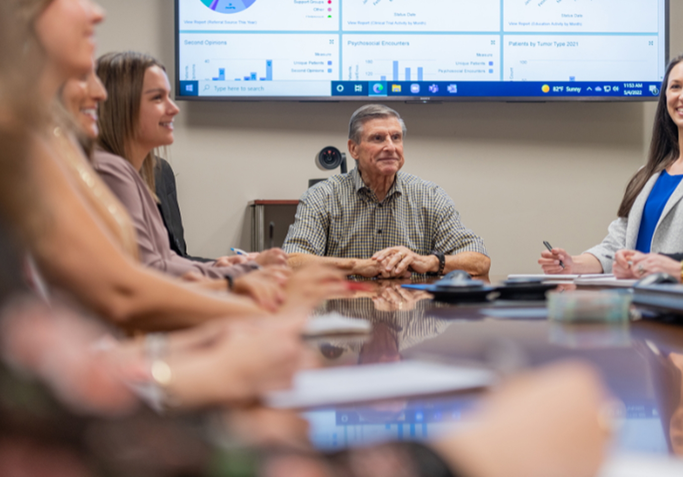 A group of people sitting around a conference table, engaged in a discussion. A presentation with charts is displayed on a screen.
