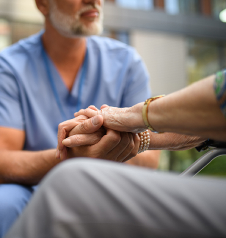 A caregiver in blue scrubs gently holds an elderly person’s hand, conveying care and compassion.