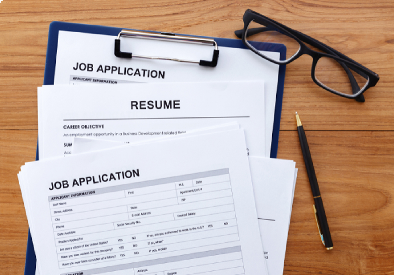 Job application and resume forms on a wooden desk, accompanied by eyeglasses and a pen, suggest a professional and organized setting.