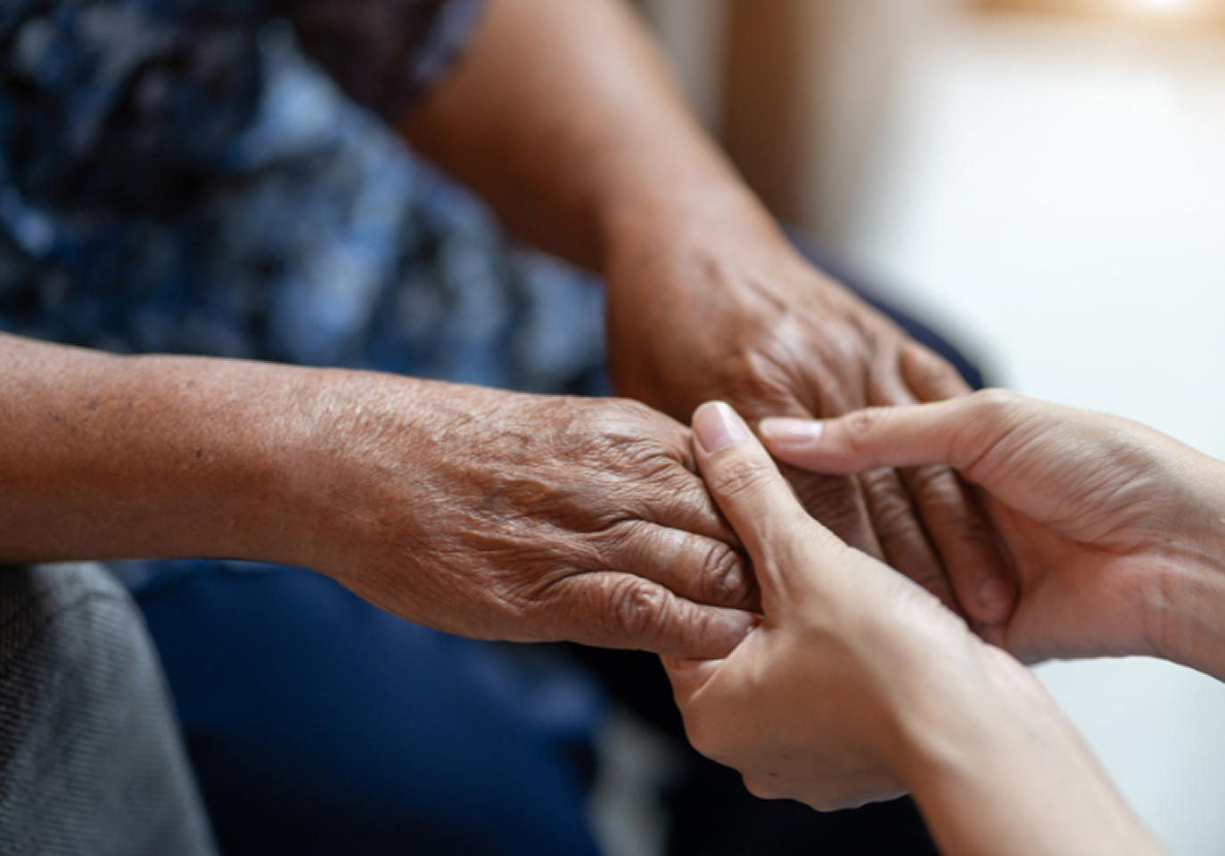 Two hands gently hold and comfort an elderly person's hands. The background is softly blurred, emphasizing the warmth and care in the interaction.
