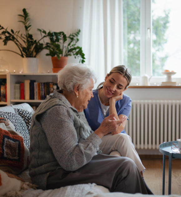 A caregiver in blue scrubs sits beside an elderly woman on a cozy couch, sharing a warm conversation. The room has plants, books, and soft lighting, creating a comforting, homely atmosphere.