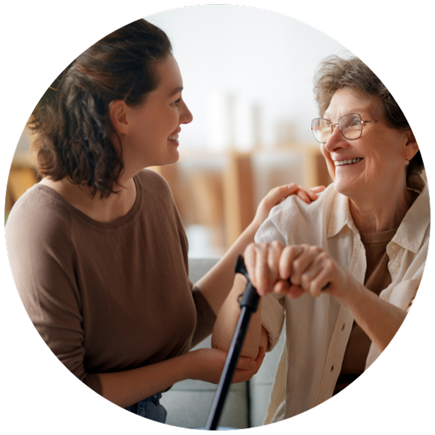 A young woman and older woman smile warmly at each other. The older woman holds a walking cane, conveying a sense of care and companionship.