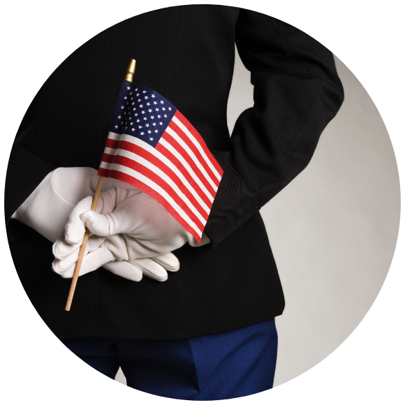 A person in a dark uniform with white gloves holds a small American flag behind their back.
