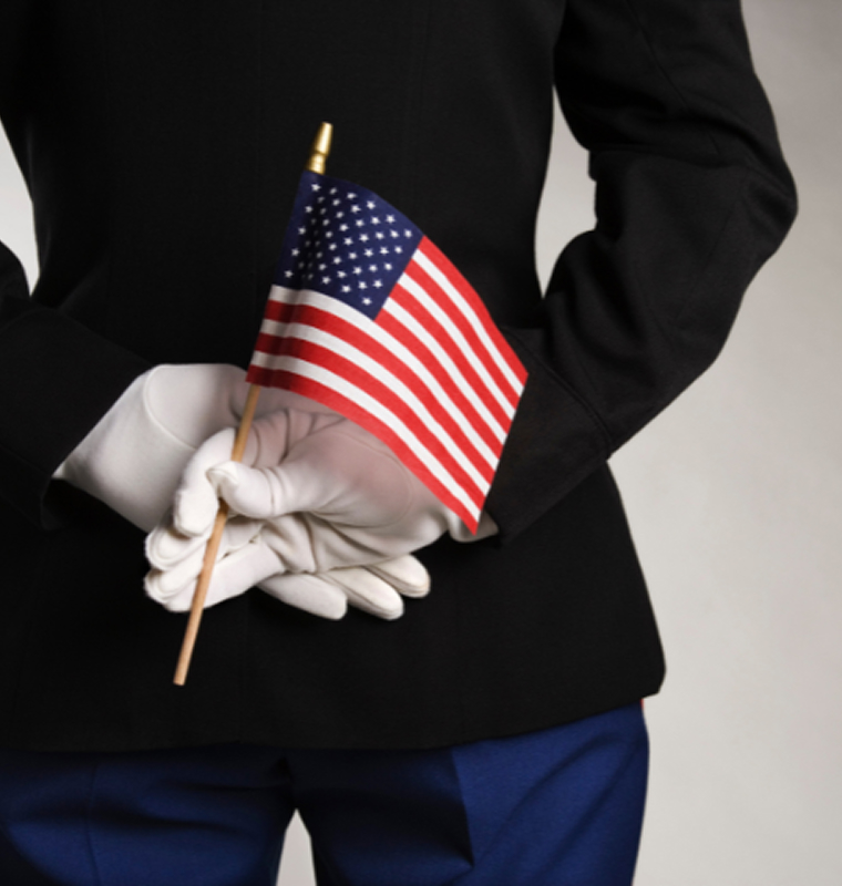 A person in a dark uniform with white gloves holds a small American flag behind their back.