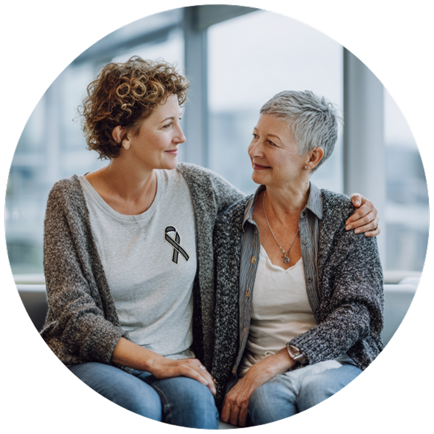 Two women sit closely on a bench indoors, sharing a warm and supportive glance. One wears a shirt with a ribbon symbol.