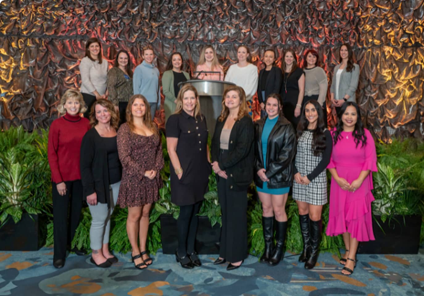 A group of women stands in two rows, smiling confidently. They're dressed in diverse, stylish outfits. The background features textured fabric and green plants, conveying a professional and celebratory atmosphere.