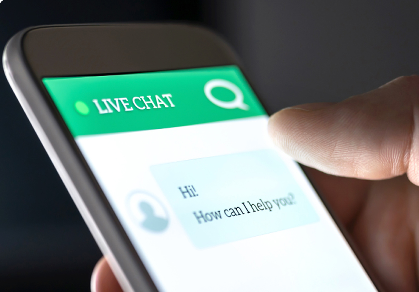 Close-up of a finger touching a smartphone screen displaying a live chat interface. The screen shows a message saying, "Hi! How can I help you?" in a chat bubble.