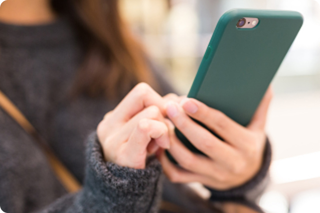 A person wearing a dark sweater uses a smartphone with a green case. The setting is softly blurred, emphasizing focus and engagement with the device.