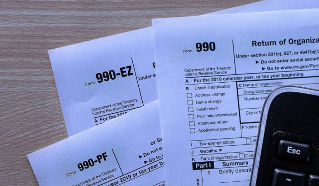 A close-up of IRS tax forms 990, 990-EZ, and 990-PF on a wooden surface. A black calculator partially visible suggests financial context.