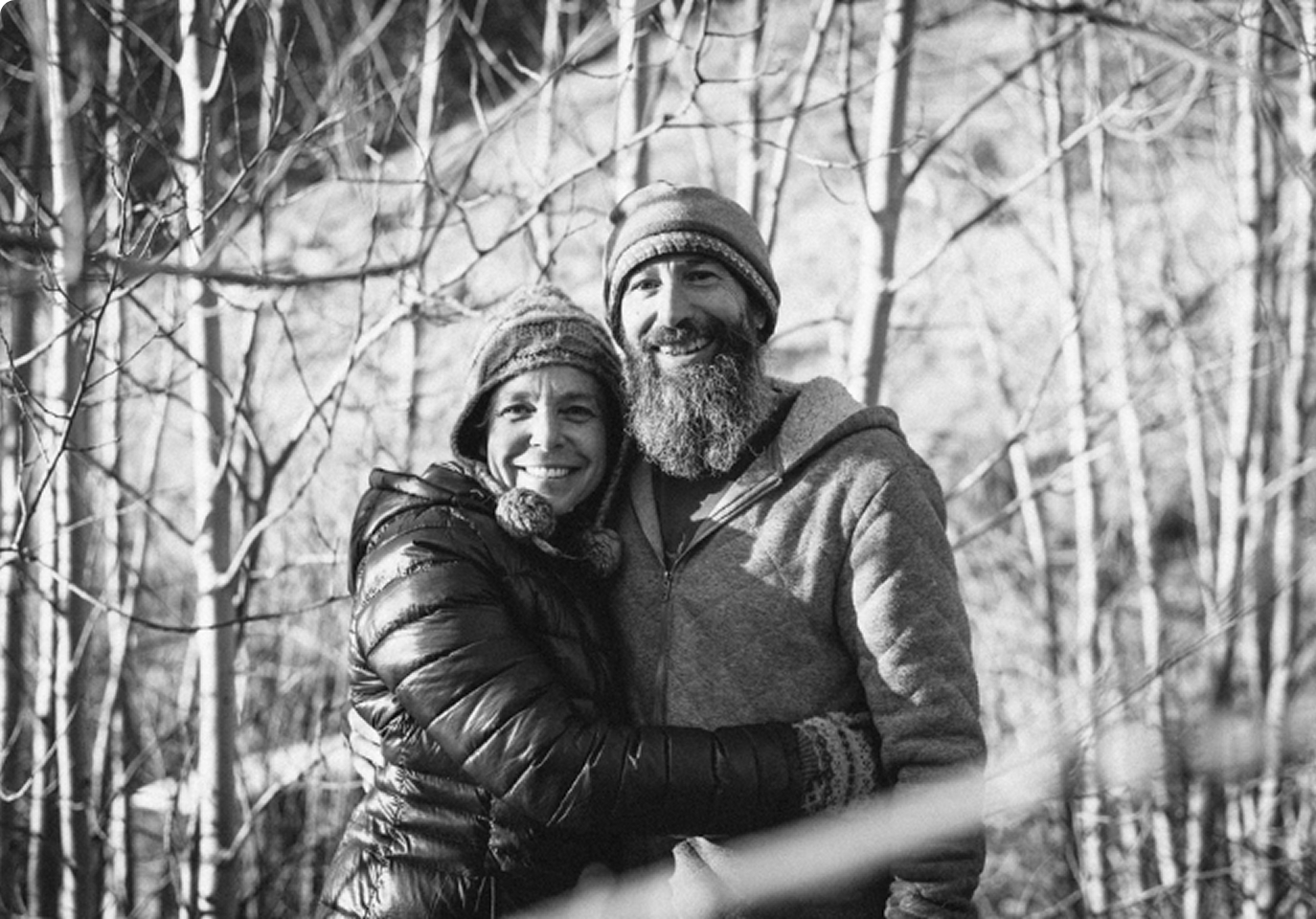 A couple in winter clothing smiles warmly, embracing in a sunlit, leafless forest.