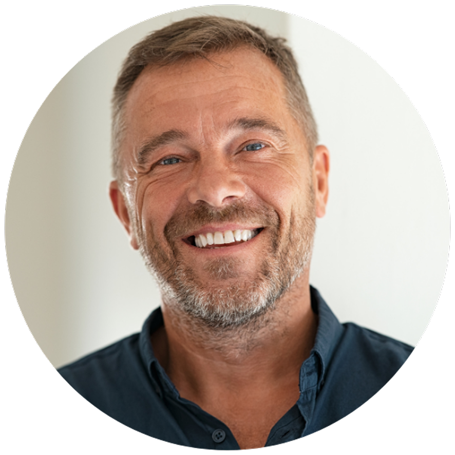 Smiling man with short hair and beard, wearing a dark blue shirt. His expression is joyful, conveying warmth and friendliness against a plain background.