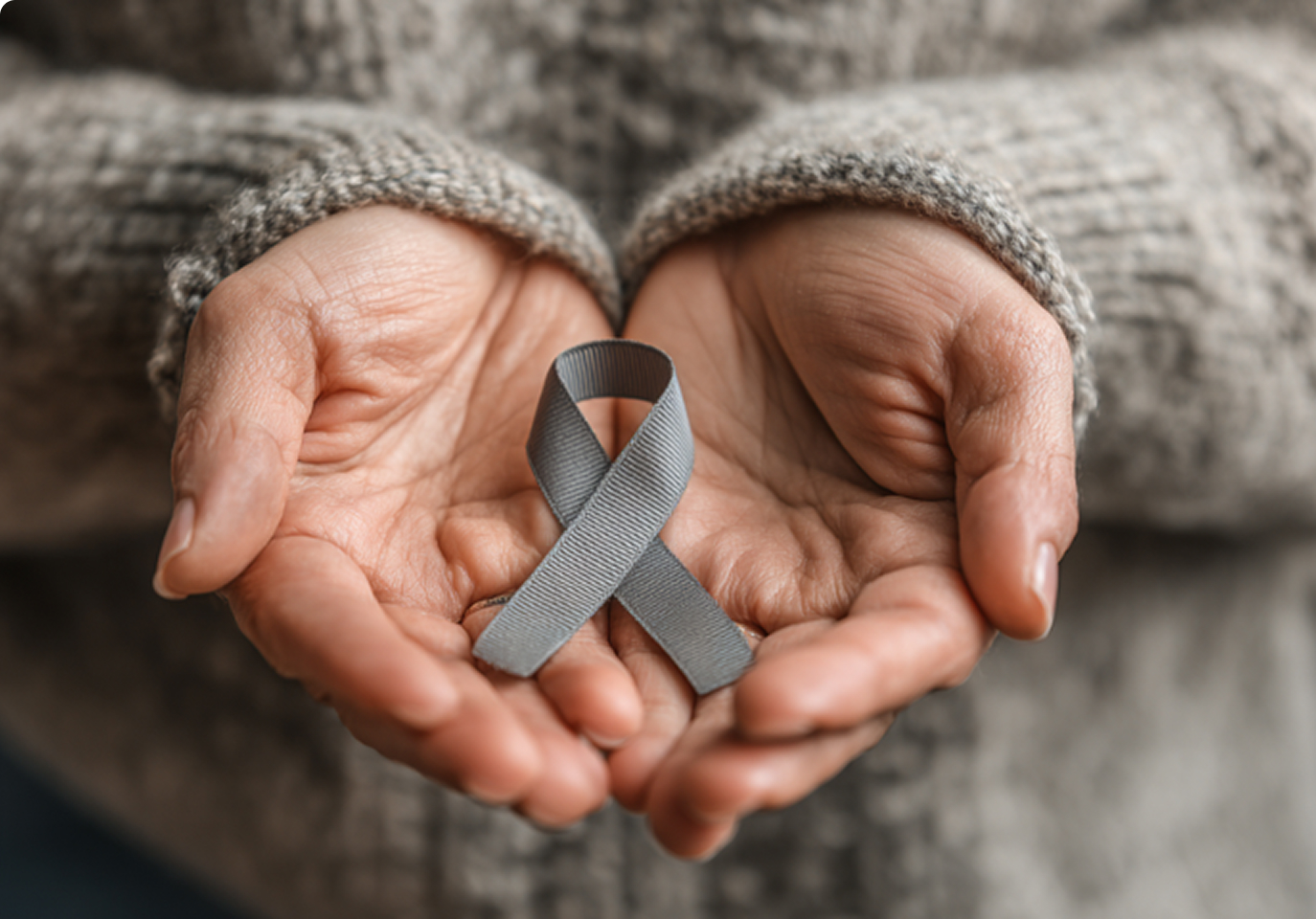 Hands gently hold a gray awareness ribbon, symbolizing support and care. The wearer's sweater sleeves are visible