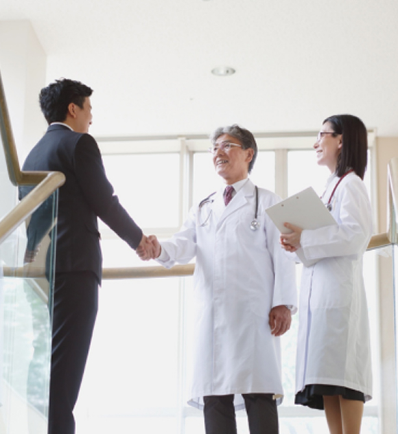 A businessman shakes hands with a smiling male doctor in a white coat, while a female doctor with a clipboard looks on.