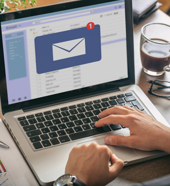 A person checks their laptop, displaying an email inbox with a new message notification. Nearby are a cup of coffee and glasses, suggesting a focused, work-like atmosphere.