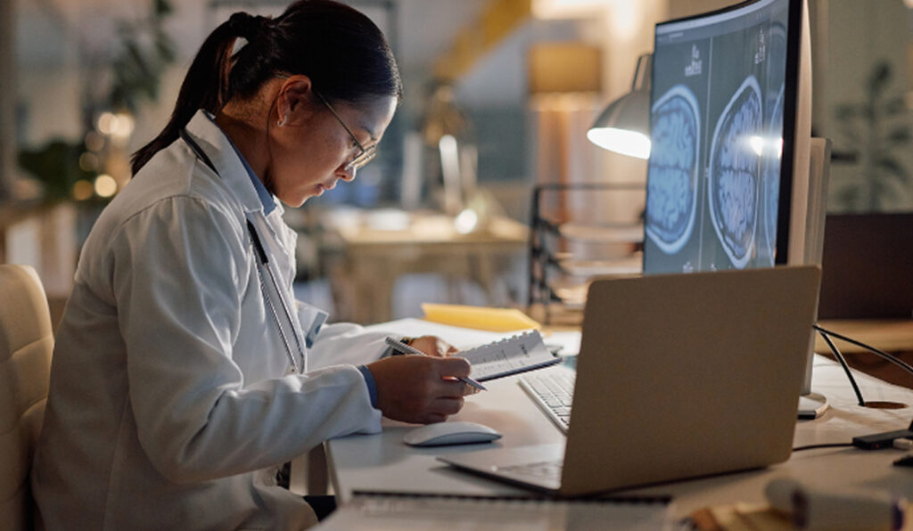 A focused doctor in a white coat reviews brain scans on a computer screen, taking notes at a desk.