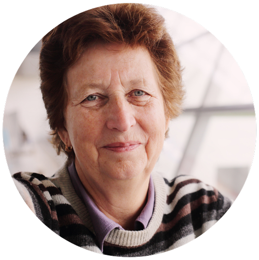 Smiling older woman with short brown hair wearing a striped sweater and purple shirt. Soft natural light and blurred neutral background.