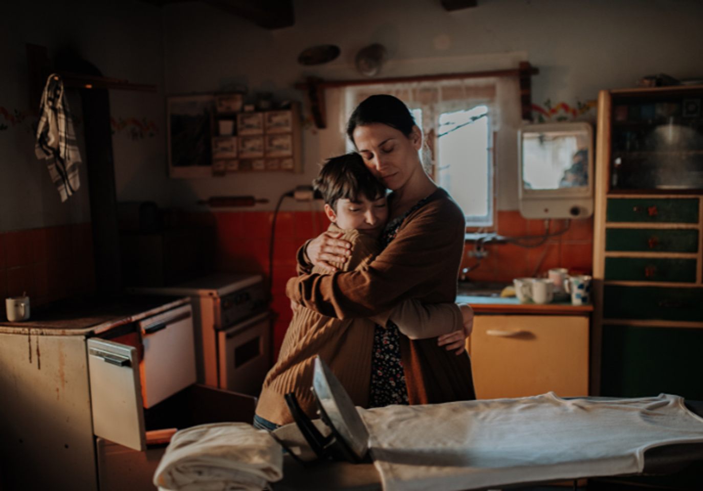 A woman gently hugs a boy in a cozy, dimly-lit kitchen, with an ironing board in the foreground.