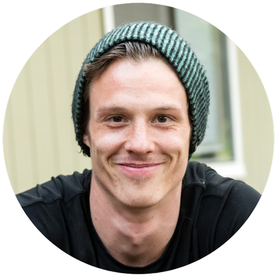Young man with a warm smile wearing a green-striped beanie and black shirt. Background shows a blurred, light-colored wall. Casual and friendly tone.