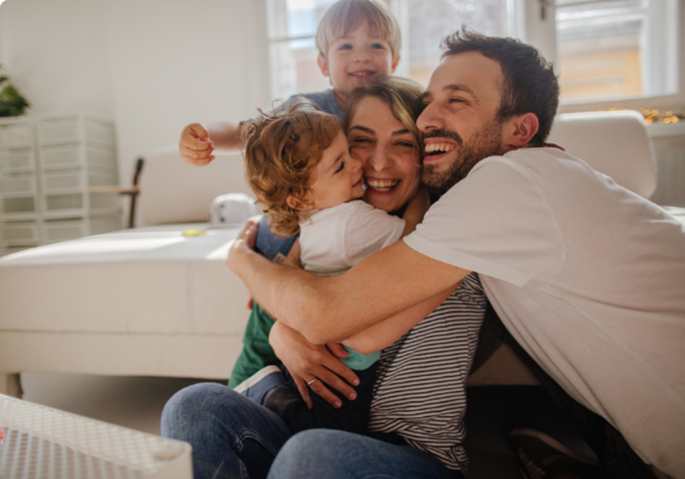 A joyful family of four embraces tightly on a couch in a bright living room. Smiling parents with two young children convey warmth and happiness.