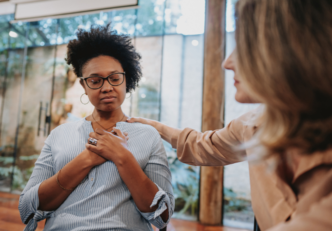 A person wearing glasses looks thoughtful, holding their chest in a serene room. Another person offers comfort with a hand on their shoulder, indicating support.