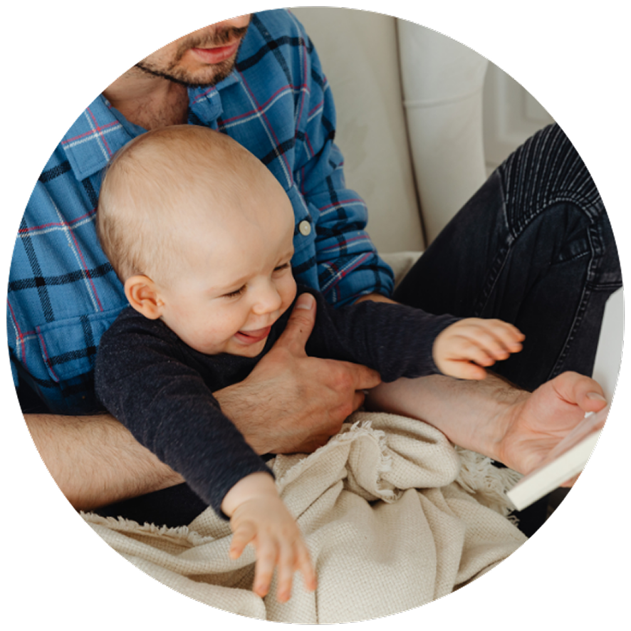 A joyful baby reaching forward, sitting on an adult's lap. The adult wears a blue plaid shirt, and they are reading together on a cozy couch.