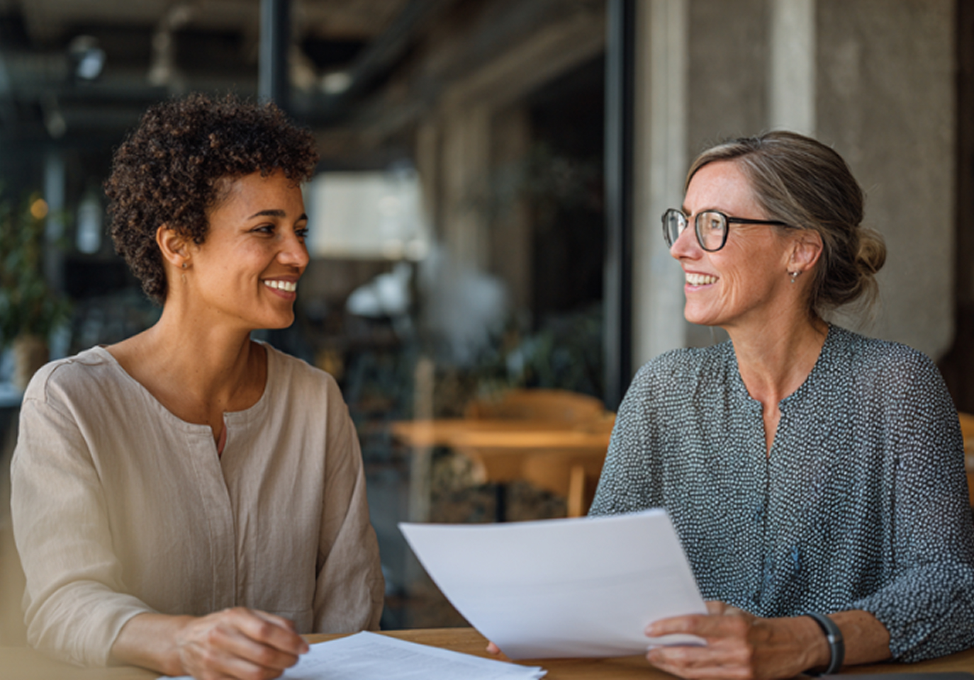 Two women sit at a table in a cafe, smiling and holding documents. The atmosphere is warm and friendly, suggesting a positive discussion.
