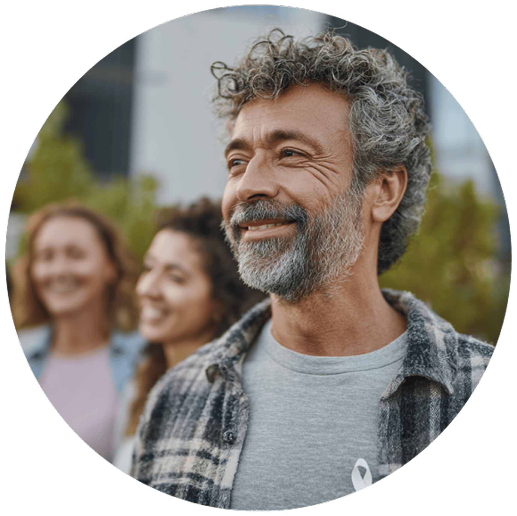 A middle-aged man with curly gray hair and a beard smiles warmly, wearing a plaid shirt and gray T-shirt. Two blurred people are in the background, conveying a joyful, outdoor setting.