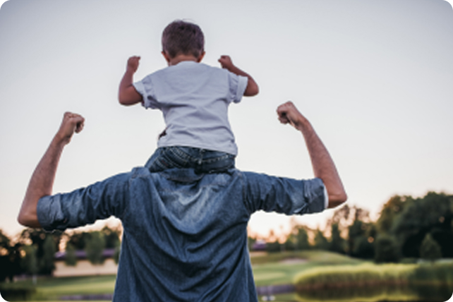 A man in a denim shirt carries a child on his shoulders, both with arms raised in a playful victorious pose.