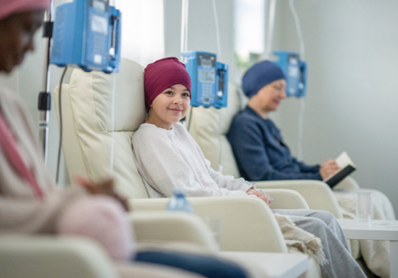 A smiling child wearing a red headscarf sits in a hospital chair receiving treatment. Two other patients, also in scarves, sit nearby; one reads a book.