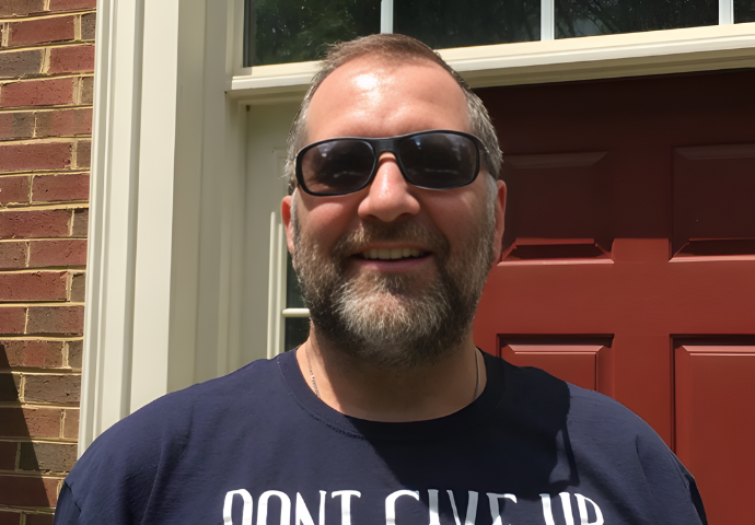 Adult man smiling outdoors while wearing sunglasses in front of his home.
