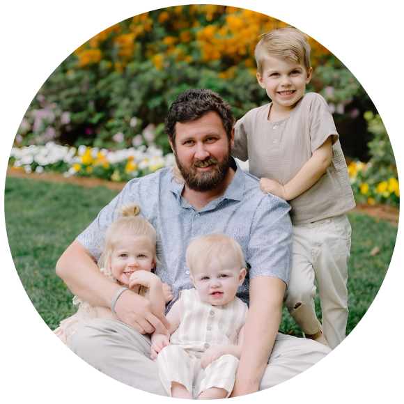Father sitting on the grass with three young children, smiling together outdoors.