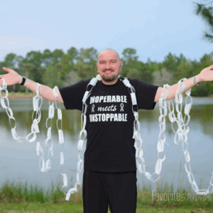 Man standing outdoors with arms open, holding paper chains by a lake.