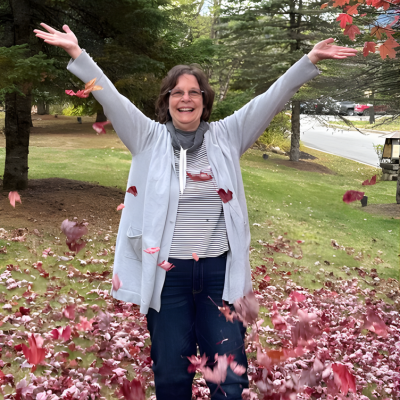 Woman outdoors with arms raised as autumn leaves fall around her.