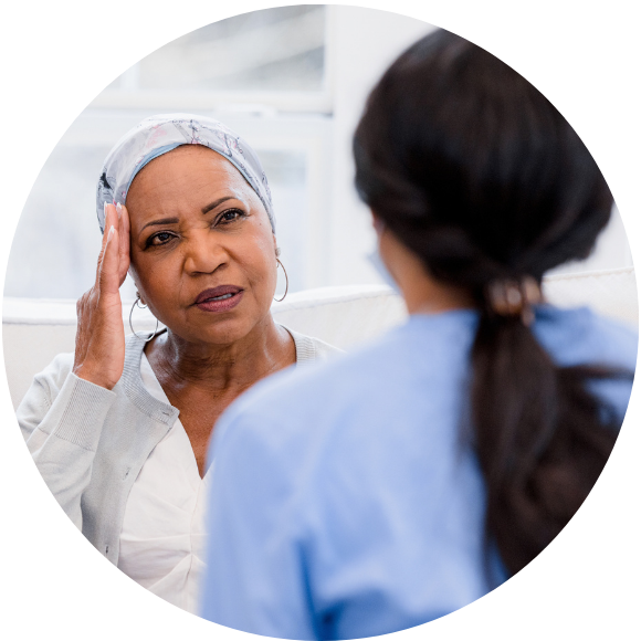 An adult woman in a head scarf gently touches her head with her hand, indicating headache. She is speaking with a female professional, likely medical, who listens intently to her patient's issue