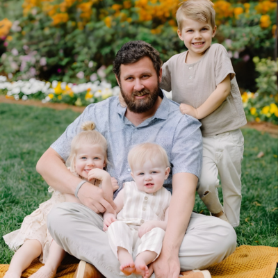 Father sitting on the grass with three young children, smiling together outdoors.
