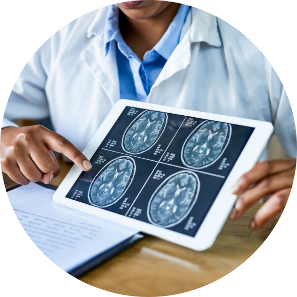 A healthcare professional in a white coat points to brain MRI images displayed on a tablet during a medical discussion, with scan details visible alongside paperwork on a desk.