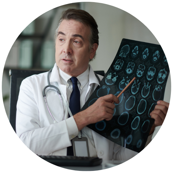 A physician in a white coat with a stethoscope holds up a sheet of brain imaging scans and points to a specific area with a pencil while explaining findings during a medical consultation.