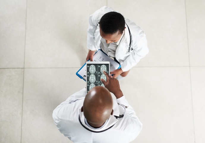 Two physicians meet in an open professional space, one has a brain MRI pulled up on an ipad, the other holds a medical chart