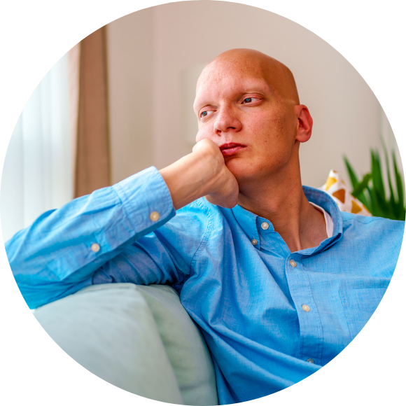 A bald adult wearing a blue button-down shirt sits on a couch, resting their head on their hand and looking off to the side. The setting is a quiet home space with soft light, bookshelves, and plants. The expression suggests reflection, fatigue, or uncertainty.