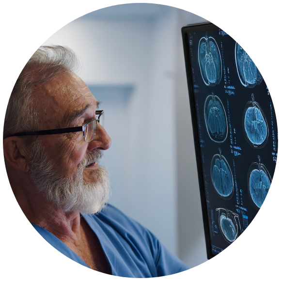 An older male healthcare professional with gray hair and a beard studies brain MRI scans displayed on a vertical monitor in a clinical setting, focused on detailed cross-sectional images of the brain.