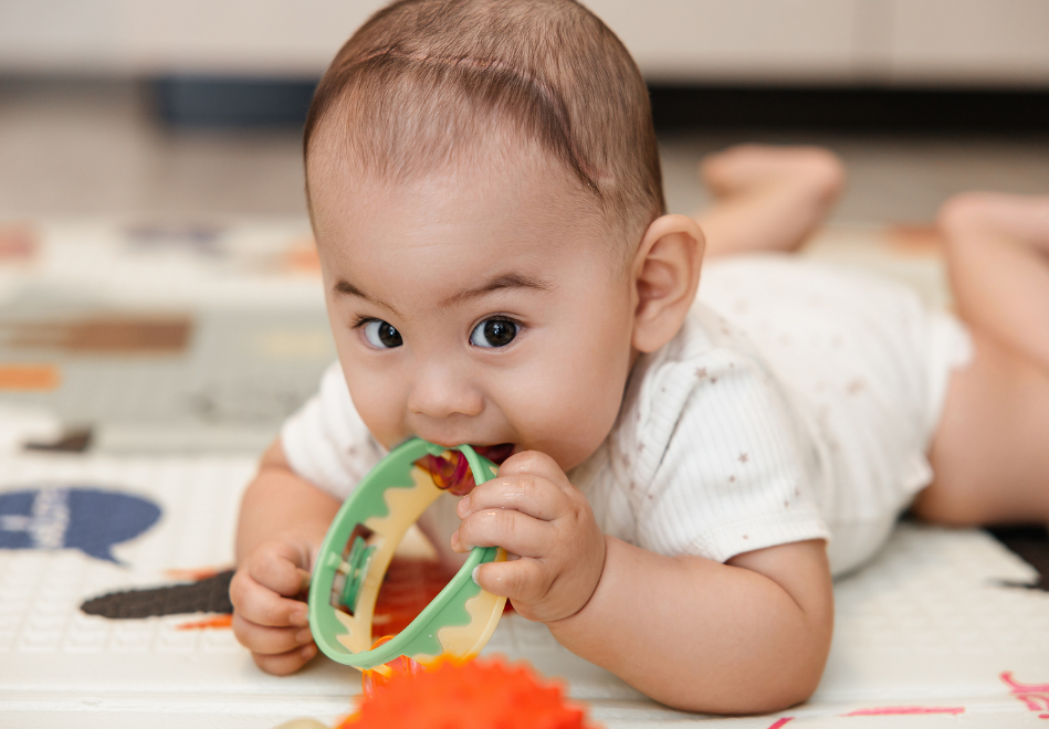 A baby with a craniotomy scar lies on their stomach on a play mat, holding and chewing on a colorful teething toy, with bright eyes focused forward in a calm indoor setting.